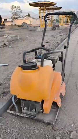 A plate compactor is being used to flatten a gravel and dirt pathway. In the background, there are yellow pavilions, stacks of paving stones, and a chain-link fence. The sky is partly cloudy.