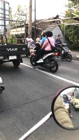 A two-wheeled vehicle is on the left side of the screen, partially in view, with a license plate that reads "VT 11-28." Next to it, a black pickup truck with the brand "VIAR" visible on the side travels on the road. On the right, a red and white motorcycle carries two passengers. The person driving the motorcycle wears a blue shirt and black pants, while the passenger in the back wears a pink shirt and black pants. They are moving in the same direction as the truck. Further to the right, a person on another motorcycle, wearing a purple outfit and a white helmet, is also visible. The environment appears to be an urban street with buildings and trees in the background, under what seems to be daylight.