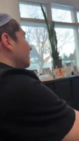 A young man wearing a dark shirt and a blue, white, and yellow kippah is filmed indoors. He is looking to the right, and then turns his head to look directly at the camera, then back to the right, as a voice repeats "Jew boy" multiple times. A window with bare trees and a snake plant in a black pot are visible in the background. A black cabinet is behind him. The natural light is bright. The video is close-up and handheld, with some slight movement.