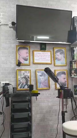 The video shows a section of a barber shop, featuring a gray brick wall decorated with six framed pictures of men's hairstyles. A flat screen television is mounted above the frames. To the left of the picture display is a rolling black cart with multiple shelves, and a hairdryer. To the right is a taller silver stand with a hairdryer attached, positioned beside a white stool. Shelves filled with hair products are visible in the background.