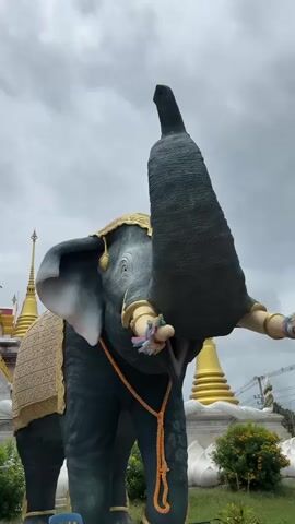 A low-angle shot captures a statue of a dark grey elephant with its trunk raised high against a cloudy sky. The elephant statue is adorned with a golden headdress and a decorated saddlecloth. Its trunk is curled and positioned upwards, and colorful tassels are tied to its tusks. In the background, a golden stupa and other ornate structures of a temple are visible, suggesting a religious or cultural setting. The overall atmosphere is overcast and slightly dramatic due to the low camera angle and the imposing presence of the statue.