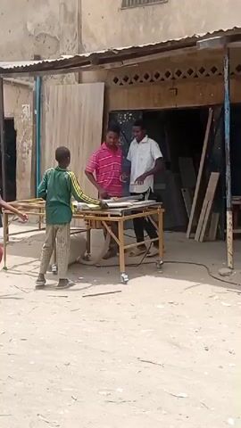 The video depicts an outdoor carpentry workshop in Gama, Nigeria, during the daytime under bright sunlight. Several young boys and at least one adult male are engaged in woodworking activities. One adult, dressed in a light blue shirt, is seen operating a power tool, possibly a drill or sander, on a piece of wood at a workbench. Other young boys are gathered around different workbenches, observing or participating in tasks related to carpentry. In the background, a partially assembled white cabinet or shelving unit is visible, along with various wood planks and materials. A boy in a blue and white striped shirt and red pants walks across the dusty ground, carrying a white object, adding to the bustling atmosphere of the open-air workshop. The setting features a simple building with an exposed brick or adobe wall and a corrugated metal roof providing shade over the work area.