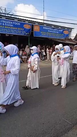 A procession of people, largely children, walks down a paved road on a clear day. The children are dressed in white robes and head coverings, some in white hats and others in blue hijabs. Several boys wear batik patterned sarongs. Onlookers, a mix of adults and children, are seated on the side of the road, watching the procession. Banners are visible above the shops lining the street, advertising frozen food and a plastic shop. A motorcycle is parked on the side of the road amongst the seated crowd. The overall scene is one of a community event or parade.