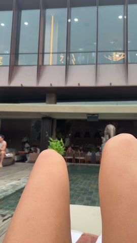 The video shows a view from a poolside perspective, initially focusing on a person's knees, then panning upwards to show a modern building with large, rectangular windows. Inside the building, people can be seen exercising on treadmills, suggesting it may be a gym. Below the gym level, there is an open-air restaurant area with people sitting at tables. Near the pool, two men are seated at a table with an umbrella. The weather appears overcast.
