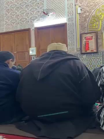 The video shows a scene inside a mosque. Several men are seated on the floor, facing forward, presumably engaged in prayer or meditation. The men are wearing various types of robes or hooded garments. One man is wearing a black hooded robe with a light-colored cap, while another is in a black and white checkered robe. The camera pans slightly from left to right, revealing more men in similar attire. The interior of the mosque is adorned with intricate tile work and ornate carvings. There are wooden doors, a digital display with red numbers, and a decorative niche with a light fixture. A small fan is visible near the ceiling. The scene appears peaceful and solemn.