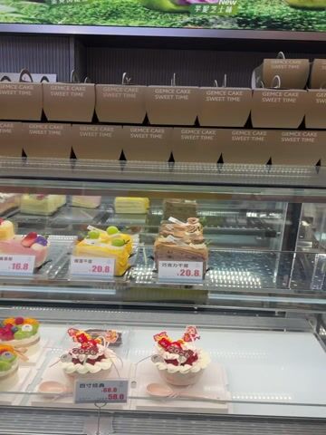 The video shows a display case filled with various cakes and pastries in what appears to be a bakery or cafe. The cakes are decorated with fresh fruit, cream, and other toppings. Price tags are visible for each item. Brown paper bags labeled "GEMCE CAKE SWEET TIME" are stacked above the display case. Some pre-packaged pastries are also visible. The environment is brightly lit.