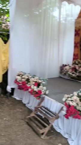 The video displays an outdoor gathering area, possibly for an event, under a large canopy. In the foreground, a white fan with a black head stands on a rolling base. The ground is covered with dirt and scattered leaves. Behind the fan, a yellow fabric is draped, possibly serving as a backdrop or partition. Rows of white chairs are set up, many covered with white fabric. Some of these chairs are placed around green tables. One red plastic chair stands out amidst the white ones. In the middle ground, a man in a black t-shirt walks away from the camera, and a woman in a green hijab sits on a chair. Further back, more chairs, some green plastic ones, are visible. The overall environment suggests a partially prepared outdoor event space.