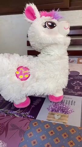 A stuffed toy llama, white with pink accents on its ears, mane, hooves, and a musical note emblem on its side, is positioned on a table covered with a patterned cloth. The llama's head and body move rapidly, and its eyes are visible. The background shows wooden furniture. The video appears to be a close-up shot of the toy.