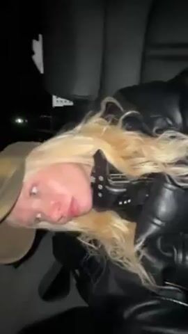 A woman with long blonde hair, pale skin, and closed eyes is reclined in what appears to be a car. She wears a dark leather jacket and a studded collar. The background is dark, suggesting it is nighttime. The car interior is visible, including parts of the seats and a vent. The woman puckers her lips slightly.