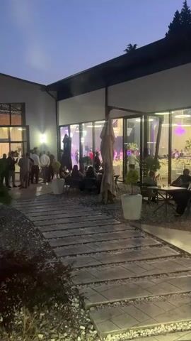 The video shows a nighttime outdoor gathering with people milling around. The scene is illuminated by artificial lights from inside a building with large glass windows, and there are also some outdoor lights. A stone pathway leads into the frame from the bottom, bordered by gravel and some sparse foliage. Several tables and chairs are set up outside, some occupied by people. Inside the building, visible through the glass, are more tables set for an event, with people seated and standing. The sky is dark blue, indicating evening or night.