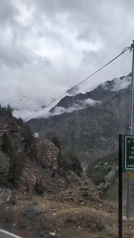 The video shows a bus traveling on a narrow, winding mountain road under a cloudy sky. The road is carved into the side of a steep, rocky mountain, with sparse vegetation. The bus appears to be the only vehicle on the road at this time. The terrain is rugged and mountainous, indicating a remote or high-altitude location. The weather appears overcast, with clouds obscuring the mountain peaks in the distance. The camera is positioned on a higher vantage point, looking down on the road and the bus.