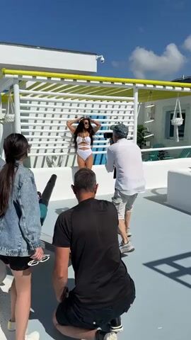 The video shows a photoshoot in progress on a rooftop or balcony. A woman in a white bikini is posing in front of a white lattice structure with yellow trim under a bright blue sky. Two men and a woman are present. One man, wearing a black t-shirt and shorts, is kneeling on the ground in the foreground, facing the woman and holding what appears to be a leaf blower. Another man, wearing a white shirt and shorts, stands behind and to the right of the woman, holding a camera and filming her. The woman on the left, wearing a denim jacket and shorts, is standing and watching the photoshoot. The environment appears to be a modern outdoor space with white structures and potted plants. The woman in the bikini is actively posing and interacting with the camera and the leaf blower.