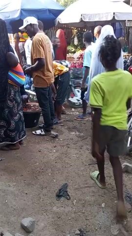 The video shows a busy marketplace scene. People are standing around stalls selling various items under blue and white umbrellas. One person in a white gown walks across the foreground. In the lower right corner, motorcycles are parked. The ground is dirt. Several people are grouped around a black bucket, possibly examining its contents.