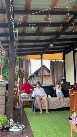 The video begins on a covered outdoor porch with a woman sitting on a couch, looking towards the camera. Two young children are also present; one is on a chair to the left, and the other is on the couch to the right. The porch is decorated with string lights and has plants and furniture. The camera then pans to reveal a larger outdoor gathering under white tents. Several people are seated at tables and on benches, suggesting a social event or party. The environment appears to be a rural or outdoor setting with trees and a wooden fence visible in the background. The sky is overcast.