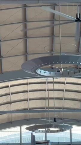 The camera is looking upwards at a high, light-colored ceiling supported by a grid of metal beams. Two large ceiling fans with multiple blades are visible, both rotating. Below the fans are two circular light fixtures, also suspended from the ceiling. The fixtures are gray and have a ring of small lights around their perimeter. The overall impression is that the video was taken in a large, modern indoor space, possibly an airport terminal or a convention center. There's a hint of a bright exterior visible through the windows or opening.