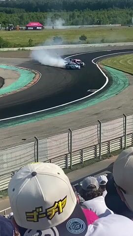 The video shows a race car drifting on a race track, creating a large amount of smoke. In the foreground, spectators are watching the event, with one person wearing a white baseball cap with Japanese writing. The race track is asphalt with green curbing and grassy areas surrounding it. In the background, there are trees and some buildings. The weather appears to be clear and sunny.