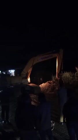 The media file shows an outdoor scene at night, illuminated by artificial lights. The primary object of focus is a large yellow excavator, identified by the "KOMATSU" logo visible on its arm. The excavator is positioned on a mound of dirt, suggesting ongoing construction or excavation work.

In the foreground, several individuals are gathered, their faces obscured by darkness or the angle of the camera. They appear to be observing the activity involving the excavator. The ground is dark and appears to be uneven, possibly a dirt road or construction site. In the background, a few lights are visible, possibly from buildings or vehicles, hinting at a nearby settlement or roadside area. The overall atmosphere is dim and somewhat mysterious due to the nighttime setting.

The time of day is clearly night, indicated by the dark sky and the reliance on artificial lighting. There are no visible cues about the weather, though the presence of water or dampness on the ground in some areas might suggest recent rain or a moist environment. The location cues, such as the excavator and the general setting, suggest an outdoor work site, possibly in a rural or semi-rural area. The presence of people watching the excavator might indicate curiosity, concern, or involvement in the ongoing task. However, without clearer facial expressions or body language, it's difficult to ascertain specific emotions.

The only visible text is the "KOMATSU" logo on the excavator. The provided location context of Bintangpermata, Indonesia, further contextualizes the scene as likely taking place in that region. The activity appears to be construction or earthmoving work being conducted at night.