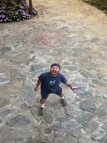 A man is standing on a stone walkway, filming with his smartphone. He has a beard and is wearing a blue t-shirt, navy shorts, and a black baseball cap. The stone walkway is decorated with a red design at its center. He looks up and smiles at the camera. The camera then shifts to show a wooden building with a thatched roof, illuminated with yellow lights. The building is surrounded by trees, and mountains are visible in the distance.
