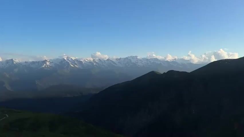 The video shows a wide drone shot of a mountain range under a clear blue sky with a few white clouds scattered across the horizon. The foreground is a dark green mountain slope with some lighter green patches. A winding dirt path is visible on the left side of the foreground. The midground consists of lush, dark green forested mountains. In the background, a majestic range of snow-capped mountains stretches across the horizon, partially obscured by atmospheric haze. The lighting suggests either early morning or late afternoon, casting shadows across the landscape. There are no people or man-made objects visible other than the path.