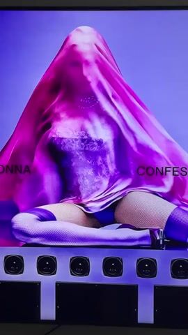 The video shows a person draped in a pink translucent fabric, obscuring most of their body. The person is wearing a purple dress with lace detailing, fishnet stockings, and what appear to be blue undergarments. Their legs are crossed, and they are seated on a dark surface. Below the seated person is a row of six speakers, each with a black circular speaker cone. The background is a gradient of purple and blue, suggesting an artificial lighting setup. Text is visible on the left and right sides of the image, reading "MADONNA" and "CONFES" respectively. The lighting is dramatic, casting shadows and highlighting the texture of the fabric and the person's attire.
