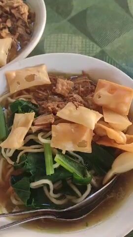 The video shows a close-up overhead view of three bowls of noodles on a green patterned tablecloth. The bowls contain noodles, broth, shredded meat, leafy green vegetables, and fried wonton pieces. Two of the bowls are closer to the camera and are nearly identical in appearance, while a third bowl is further back and slightly out of focus, but still clearly visible. The lighting appears to be natural, possibly from a window or overhead light. No people are visible in the video. The focus is entirely on the food.