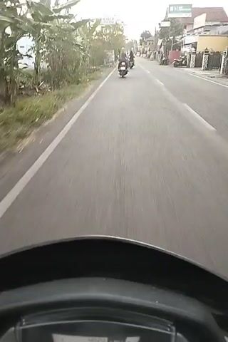 The video is shot from the perspective of someone riding a motorcycle. The rider is on a paved road with a white line marking the lane. On the left side of the road, there are trees and some vegetation. On the right side of the road, there are buildings and signs. Several other motorcycles are visible on the road, moving in the same direction as the rider. The sky is visible and appears to be overcast or hazy. The video captures the motion of the motorcycle and the passing scenery. The dashboard of the motorcycle is visible at the bottom of the screen, showing a speed of 37.