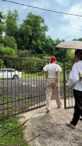 A group of people are gathered near a rusty metal gate. Two women and a person holding an umbrella are in the foreground, walking towards the gate. The woman on the left has on a white shirt with floral embroidery on the back and dark pants. The woman in the middle is holding a beige umbrella. The woman on the right is wearing a black dress and carrying a book. Behind them, two men are visible. One man in a white shirt is opening the gate, while another man in a dark shirt is walking through it. A white car is parked on the other side of the gate. The environment appears to be outdoors, with lush green trees and a cloudy sky.