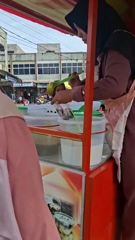 This video shows a street food stall with two women. The woman in the foreground, wearing a black hijab and a pink apron, is serving a customer. She is holding a plastic bag with food items and a serving utensil, and appears to be either preparing an order or showing the customer the available options. In the background, another woman wearing a black hijab is visible, looking towards the stall. The stall itself is red and appears to have several containers of drinks or ingredients. The environment looks like an outdoor market or street with buildings in the background. The overall scene depicts a commercial transaction at a food stall.