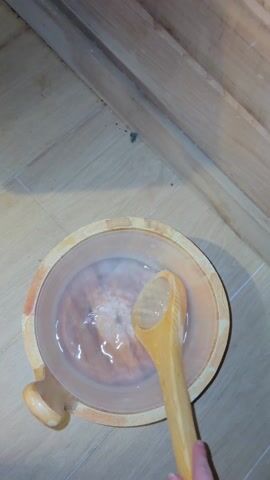 The video shows someone adding water to sauna rocks. The camera is at a high angle, looking down at a wooden bucket containing water. A wooden ladle is in the bucket. The camera moves up and right, showing a red sauna stove with a grill holding rocks. The person uses the ladle to pour water onto the hot rocks. The rocks sizzle as the water hits them. The stove has a "Finlândia Sauna" sticker on it. The sauna room is paneled with wood.