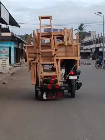 A motorized tricycle loaded with wooden furniture, including chairs and what appears to be a ladder, travels down a paved road. The tricycle is being driven by a person wearing a dark shirt and pants. Another person on a motorcycle is visible in the background to the right of the frame. The surroundings suggest a commercial area with buildings and shops lining the street. The sky is overcast, indicating a cloudy day. The tricycle has a license plate that is partially obscured. There are cylindrical objects, possibly decorative or structural, attached to the rear of the tricycle.