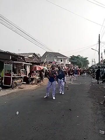 A group of people in marching band uniforms are walking down a street. The person in the foreground is wearing a blue jacket with gold trim and white pants, and is carrying a rifle with a red flag attached. They are walking in a line with other people in similar uniforms. The street is lined with buildings and stalls, and there are power lines overhead. The sky is overcast.