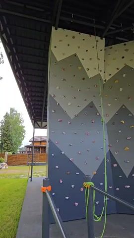 The media file shows a climbing wall with a mountainous design. The wall is constructed of several panels, and the design features varying shades of gray and white to depict mountain peaks. Numerous climbing holds in various colors, including pink, yellow, and blue, are attached to the wall. A bright green climbing rope is visible, hanging from the top of the wall and secured near the bottom. The wall is situated outdoors under a dark, industrial-style roof structure, suggesting it might be part of an adventure park or recreational facility.

In the background, to the left, a grassy area with trees and a wooden house with a shingled roof can be seen. The house appears to be a traditional Russian dacha or rural dwelling. The sky is overcast, indicating potentially cloudy weather. The scene appears to be set during the daytime. No people are visible in the media file. The overall impression is one of an outdoor recreational space designed for climbing activities. The presence of the roof suggests protection from the elements, allowing for climbing even in less than ideal weather conditions.