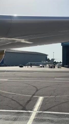 This media file captures a scene at an airport, likely a private jet terminal, given the presence of sleek, smaller aircraft. The viewpoint is from under the wing of a larger aircraft, providing a unique perspective.

In the mid-ground, a white private jet is parked on the tarmac. Several vehicles, including what appears to be a dark SUV and possibly a service cart, are near the jet, suggesting active preparation or servicing. Another smaller aircraft is visible further in the background, near a large hangar. The hangar doors are partially open, revealing more of the aircraft inside. The overall setting is an airport tarmac with paved surfaces marked with yellow and white lines, and tire tracks visible. The background shows a large, grey industrial building, likely a hangar or airport facility, and trees in the distance.

The activity appears to be related to aircraft operations, possibly pre-flight checks, boarding, or post-flight servicing. The light suggests it is daytime, with the sun casting strong highlights on the metallic surfaces of the aircraft and the wing in the foreground. The sky is bright and appears clear, indicating fair weather.

There is no visible text in the media file. The mood is professional and functional, indicative of airport operations. The scene is set in Luton, United Kingdom, implying an international airport environment.