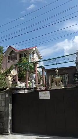 The video shows a two-story house with a red roof and white walls. There is a black metal gate in front of the house. The gate is decorated with ornamental carvings. Above the gate, there is a sign with the inscription "Въезд" and "Задерживать". The house is surrounded by greenery and trees. There are several decorative vases and statues in the yard. In the foreground, there is a stone wall and a car. The sky is blue with some clouds.