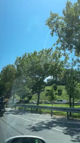 A video taken from a vehicle shows a clear blue sky. Trees line a roadside, with a grassy hill in the background. Several vehicles, including trucks and cars, are visible on the road and the hill. In the distance, a building resembling a castle can be seen. The video appears to be filmed on a sunny day.