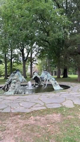 A video shows a fountain with a sculptural element in a park-like setting. The sculpture appears to be made of metal and has a greenish patina, resembling abstract figures or creatures. Water is spraying from the center of the fountain. The fountain is surrounded by flagstone paving, and behind it are large trees with green foliage. In the distance, a building with a light-colored facade and a darker roof is visible through the trees. The ground is covered with dirt and fallen leaves. The video is filmed from a slightly low angle, looking up at the fountain and the surrounding environment.