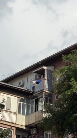 The video shows a close-up view of the side of a multi-story building against a cloudy sky. Laundry is hanging on a clothesline on a balcony. Air conditioning units are visible on the exterior walls of the building. Trees partially obscure the view on the right side. The lighting suggests it is daytime, possibly overcast.