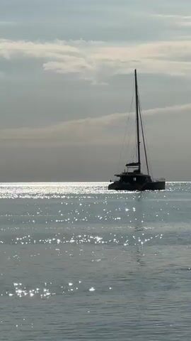 The video captures a serene marine scene with a large catamaran sailboat as the central subject. The catamaran, dark in silhouette against the bright sky, is anchored or slowly moving on calm, sparkling water. Sunlight glints intensely across the surface of the sea, creating shimmering reflections. In the background, the sky is partially cloudy, with soft, diffused light. Towards the end of the video, a dark, rocky cliff or coastline becomes visible on the right side of the frame, hinting at the proximity to land. The overall impression is one of tranquility and natural beauty, likely at morning or late afternoon given the quality of light. No people or visible text are present in the video.