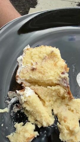 A close-up shot displays a slice of cake on a black plate. The cake is light yellow with white frosting along one edge, and appears to contain some sort of fruit filling. A spoon rests on the plate, partially covering the cake. The plate is held by a person, as indicated by a small portion of their arm visible on the left. The background includes a glimpse of a concrete surface and some greenery. The focus is on the cake and spoon, capturing their texture and details.