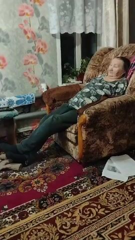 The video shows an elderly woman resting in a patterned armchair in a room with patterned wallpaper and a colorful rug. The woman is lying back with her legs extended, wearing dark pants, dark socks, and a patterned shirt. A window is visible behind her to the right, and a small table with items on it is to her left. There is a newspaper or papers on the floor near the chair.