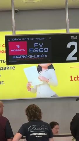 The video shows a digital display board above a check-in counter. The display board shows "CHECK-IN DESK 2" at the top. Below that, it shows the Rossiya airline logo and "FV 5969". Underneath the flight number, it says "SHARM EL SHE" and then "CHECK-IN ENDS AT 19:30" with a date of "11.08.2020". To the right of the flight information, a large "2" is displayed. Behind the display board, a cartoon of a person holding a document is visible against a yellow background. In the foreground, the backs of several people are visible, standing in a queue. One person in the center is wearing a dark t-shirt with "London" and "LEE COOPER ORIGINALS" written on it. The environment appears to be an airport check-in area.