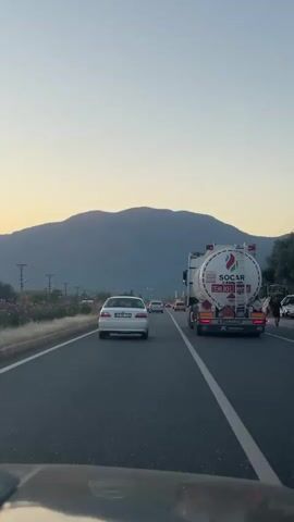 The video shows a view from inside a car, looking out the windshield onto a highway. The road has at least two lanes heading in the same direction. A white car is in the lane ahead of the camera car. To the right of the white car is a large, white tanker truck labeled SOCAR. The word "TEHLIKELI MADDE" is visible on the back of the tanker, which translates to "Dangerous Material" in English. In the background, a large, dark mountain is visible against a pale blue sky, suggesting either dawn or dusk. Other vehicles are seen further ahead in the same lane, indicating moderate traffic. The focus is slightly soft, particularly on the distant objects.