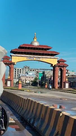 The video shows a wide shot of the Lumbini Gate, a large, ornate archway with traditional architecture and a golden dome. The gate is yellow with red trim and has "LUMBANI GATE" written on it. Two brick pillars with smaller roof structures flank the arch. A traffic sign shaped like a triangle with a cross inside is prominently displayed in the foreground, to the left of the gate. In the background, there are buildings and a clear blue sky. A person on a motorcycle is visible riding through the gate. The road in front of the gate has yellow and black striped barriers. The scene appears to be an outdoor, urban environment.