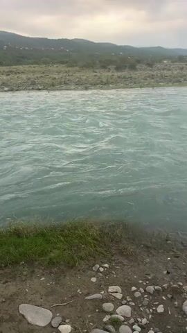 The video shows a wide, turquoise river flowing rapidly. The riverbank is lined with green grass and scattered rocks. In the background, a hilly landscape is visible, covered with trees and a few scattered buildings. The sky is overcast with gray clouds. The camera pans slowly from right to left, capturing the movement of the water and the surrounding scenery.