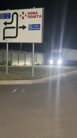 A white semi-truck with its headlights on is shown at night at an intersection. A road sign with arrows and destination names such as "Odesa" and "Kyiv" is visible above the truck. A white barrier gate with red and white stripes is partially lowered, obstructing the truck's path. The truck appears to be stopped or slowing down. The environment is dark, with some lighting from the truck's headlights and possibly streetlights. A concrete fence is visible in the background.