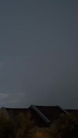 This media file captures a nighttime scene in Trap, Montenegro. The sky is dark and overcast, suggesting a cloudy or potentially stormy evening. The overall mood is somber and tranquil.

In the foreground, lush green foliage, likely trees or bushes, is visible, partially obscuring the view of what lies behind. Peeking through the greenery are the tiled roofs of several houses, their triangular gables prominently displayed. The architecture suggests a residential area. Beyond the roofs, a lighter-colored building with windows is visible on the right side of the frame, indicating the presence of more structures.

At the bottom of the frame, the silhouettes of two parked cars are discernible. One car is dark-colored, and another appears to be blue. The presence of streetlights, indicated by the warm glow illuminating the foliage and the cars, confirms that this is a developed area, likely a residential street or parking lot.

The time of day is clearly night, as evidenced by the dark sky and artificial lighting. The weather appears to be still, with no visible signs of rain or strong winds, despite the dark clouds. The overall impression is one of a quiet evening in a Mediterranean town. There is no visible text in the media file.