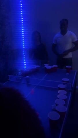 The scene is illuminated by blue LED lighting. A beer pong table occupies the center, set up for a game. Three individuals are present, one woman in a black top and two men, one wearing a white V-neck shirt. One man is holding a red cup. The atmosphere suggests a casual gathering or party setting.