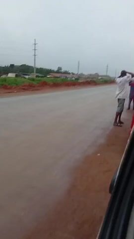 The video shows a blurry, overcast scene of a dirt road under construction. In the foreground, the edge of a dark vehicle is visible. The road itself is a reddish-brown dirt, appearing wet. On the right side of the road, a person wearing a white shirt and dark shorts is standing. In the background, there are power lines, some sparse green vegetation, and what appear to be buildings under construction or newly built. Further down the road, several motorcycles are visible, some with riders. The overall impression is of a developing area with ongoing road work.