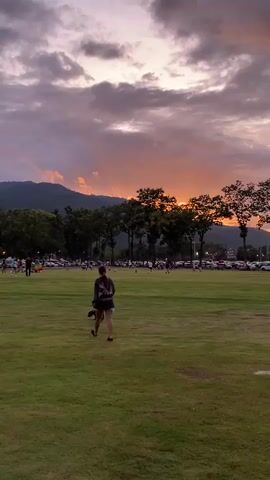 A young woman with dark hair, wearing a dark t-shirt and shorts, walks across a grassy field with a small dog on a leash. The dog, brown and white, is walking beside her. In the background, there are trees, a line of parked cars, and people walking. Mountains form a backdrop against a cloudy sky with a sunset in progress, casting an orange glow. The time is evening.