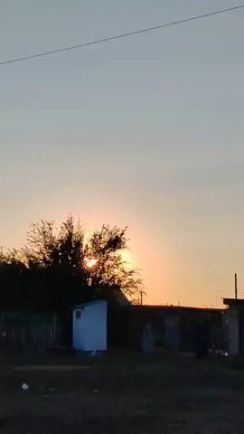 The video begins with a view of a pale blue sky with a thin wire stretching across it. The sun is beginning to set, casting a warm glow on the horizon, and silhouetting a line of trees and some buildings. As the video progresses, the camera pans slightly, revealing more of the rural landscape. There are some small, dilapidated structures, overgrown vegetation, and a rusty piece of farm equipment in the foreground. The sky remains visible throughout, transitioning from a light blue to a softer, more muted hue as the sun dips lower. The audio consists of a voice repeating the word "sky" multiple times, in Russian.